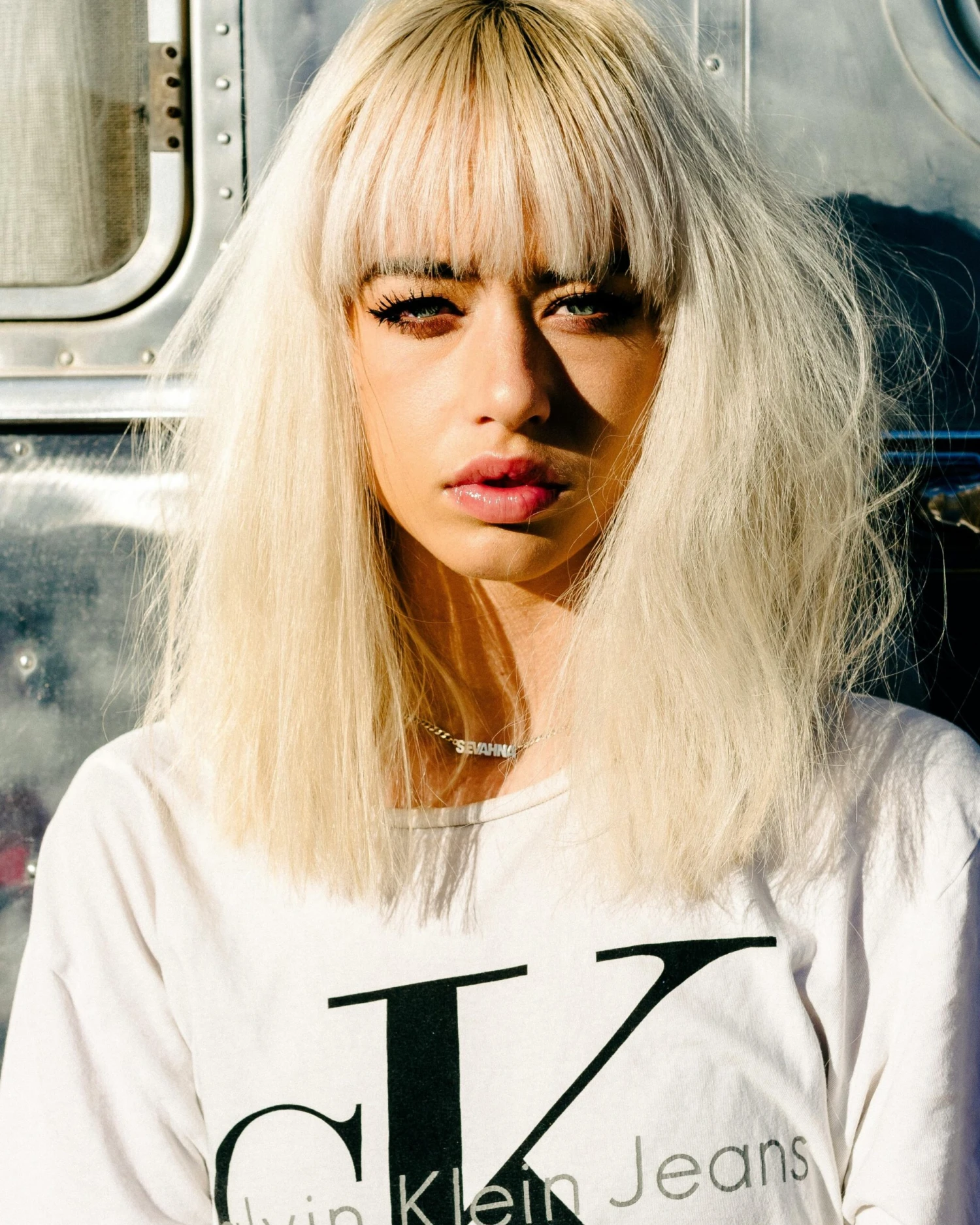 Close-up portrait of a blonde woman with bangs, wearing a white t-shirt and standing outdoors in daylight.